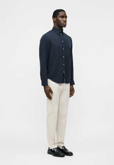 Mężczyzna stojący na tle jednolitego białego tła, ubrany w ciemnoniebieską koszulę zapinaną na guziki, beżowe spodnie i czarne eleganckie buty.