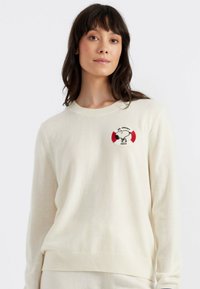 Crème gebreide trui met een ronde halslijn, voorzien van een geborduurd logo van een rode en witte reddingsboei. Zachte textuur en lange mouwen.