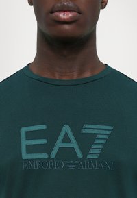 Zöld pamut póló, amelyen hímzett "EA7 Emporio Armani" logó található textúrázott részletekkel és kerek nyakkivágással.