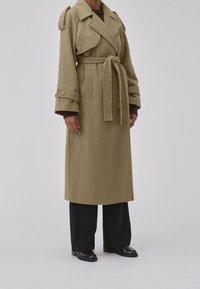 Kahvinvärinen trenssitakki, jossa on vyötärövyö, leveät kaulukset ja napilliset hihansuut. Valmistettu teksturoidusta kankaasta, A-linjaisella siluetilla ja polvenpituudella.