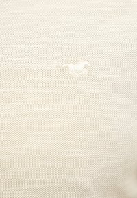 Tissu blanc texturé avec un petit logo brodé d’un cheval au galop dans une teinte légèrement plus claire.