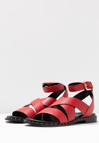 Punaiset nahka sandaalit, joissa on ristikkäin hihnat, nilkkasolki, musta pohja, kultaiset niitit reunassa, litteä muotoilu, avonainen varvas.