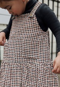 Vestito quadrettato rosa e marrone con tasca, bretelle con bottoni, orlo arricciato e texture morbida, indossato sopra una maglia scura.