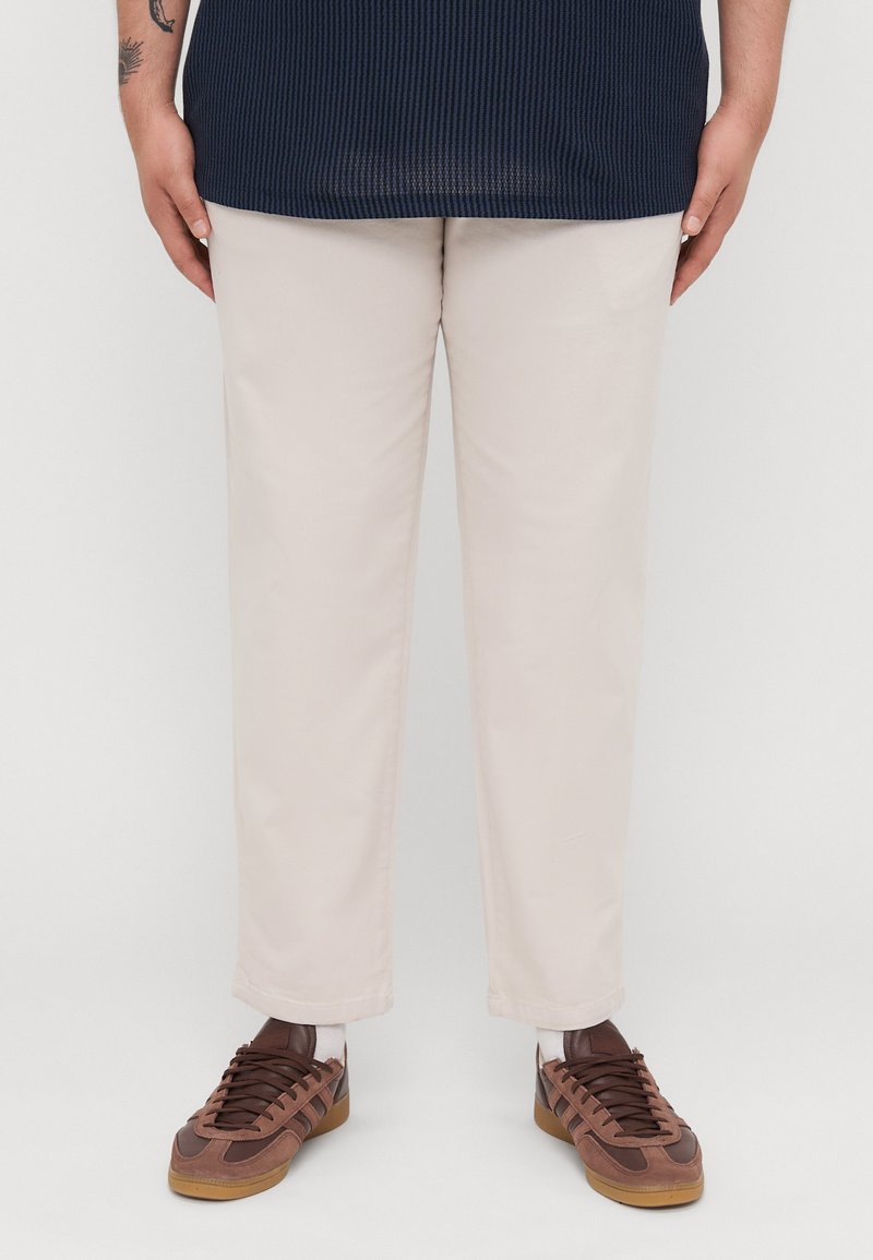 Beige straight-leg trousers with a smooth texture, paired with brown sneakers featuring a mesh upper and laces, standing on a white background.