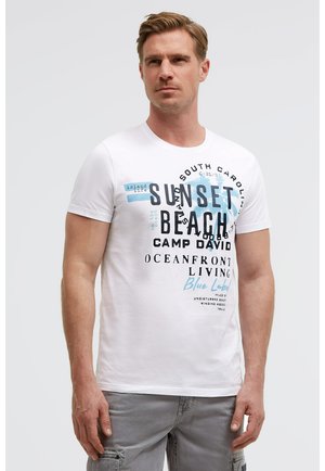 Mann trägt weißes T-Shirt mit blauem und schwarzem Text "Sunset Beach Camp David Oceanfront Living" und graue Cargo-Hose, schaut nach vorne.