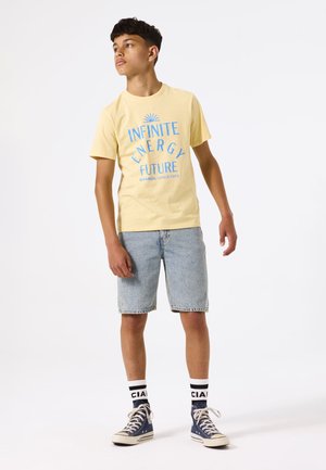 T-shirt en coton jaune avec un texte graphique bleu "ÉNERGIE INFINIE FUTUR." Portée avec un short en denim clair et des baskets montantes navy.