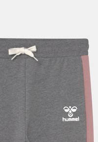 Pantalons de survêtement gris avec un cordon blanc, présentant un panneau latéral rose et un logo blanc sur le bas de la jambe. Texture en coton doux.