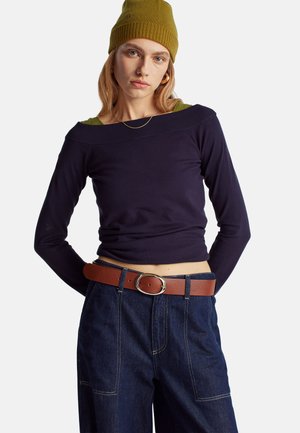 Maglione navy a maniche lunghe con design senza spalle, abbinato a una maglietta verde sotto. Cintura in pelle marrone con fibbia argentata sopra jeans in denim scuro a gamba larga.