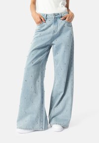 Osoba u svijetloplavim širokim trapericama ukrašenim raspršenim malim srebrnim zakovicama, bijelim tenisicama i uvučenoj bijeloj majici.