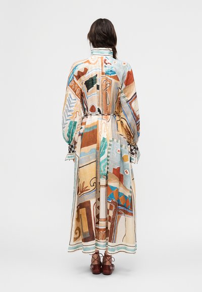 Γυναίκα που φοράει μακρύ, πολύχρωμο φόρεμα με αφηρημένο σχέδιο και φουσκωτά μανίκια, καθώς και καφέ δερμάτινα παπούτσια με κορδόνια, με την πλάτη προς το μέρος μας σε απλό φόντο.