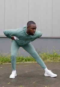 Atletický outfit v světle zelené látce, skládající se z mikiny na zip a přiléhavých kalhot, kterým dominují bílé tenisky. Na kalhotách je výrazný textový detail.