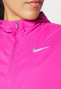 Chaqueta deportiva rosa con cuello con cremallera, hecha de tela ligera, que presenta un logo plateado de Nike en el pecho y una superficie texturizada.