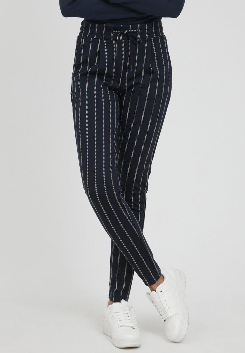 Pantalon rayé marine avec taille élastique et cordon de serrage. Coupe slim avec des rayures verticales blanches. Porté avec des baskets blanches.