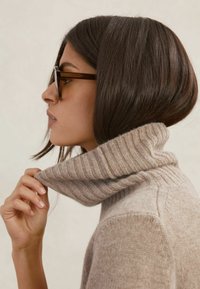 Béžový rolákový sveter s žíhaným golierom, mäkkou textúrou a priliehavým strihom. Má vysoký golier a čiastočne ho drží ruka.