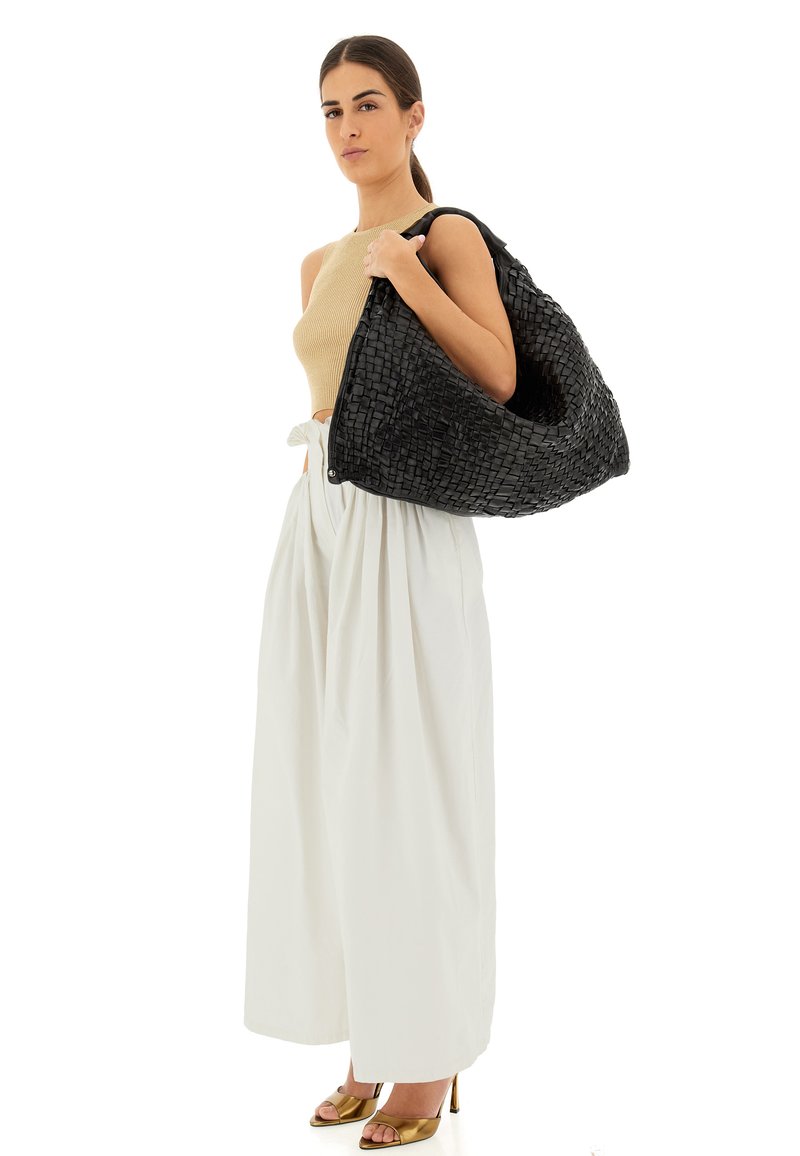 Borsa nera intrecciata a forma di hobo, con superficie testurizzata, ampia apertura e manico robusto. Il modello indossa una maglietta beige e pantaloni bianchi.