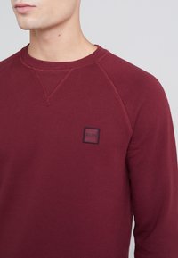 Sudadera burdeos con cuello redondo, mangas raglán y un pequeño parche de logo cuadrado negro en el pecho. Textura de tela de algodón suave.
