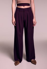 Pantaloni a gamba larga in tessuto viola intenso, con pieghe e fronte piatto, abbinati a ballerine marroni a punta. Tessuto liscio, design moderno.