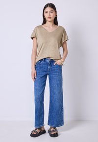 Beige kurzärmeliges V-Ausschnitt-Oberteil kombiniert mit blauen weit geschnittenen Jeans. Das Outfit wird durch schwarze Sandalen mit dekorativer Hardware ergänzt.