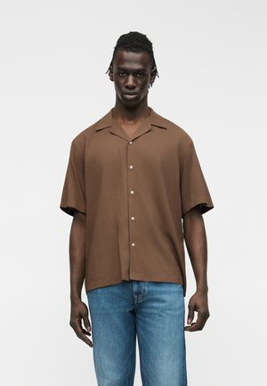 Homme à la peau foncée portant une chemise ample marron à manches courtes et un jean bleu clair, sur un fond blanc uni.