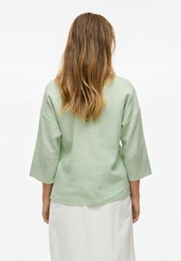 Blusa verde chiaro a maniche lunghe con tessuto strutturato, vestibilità ampia e maniche larghe, mostrata da dietro su uno sfondo bianco.