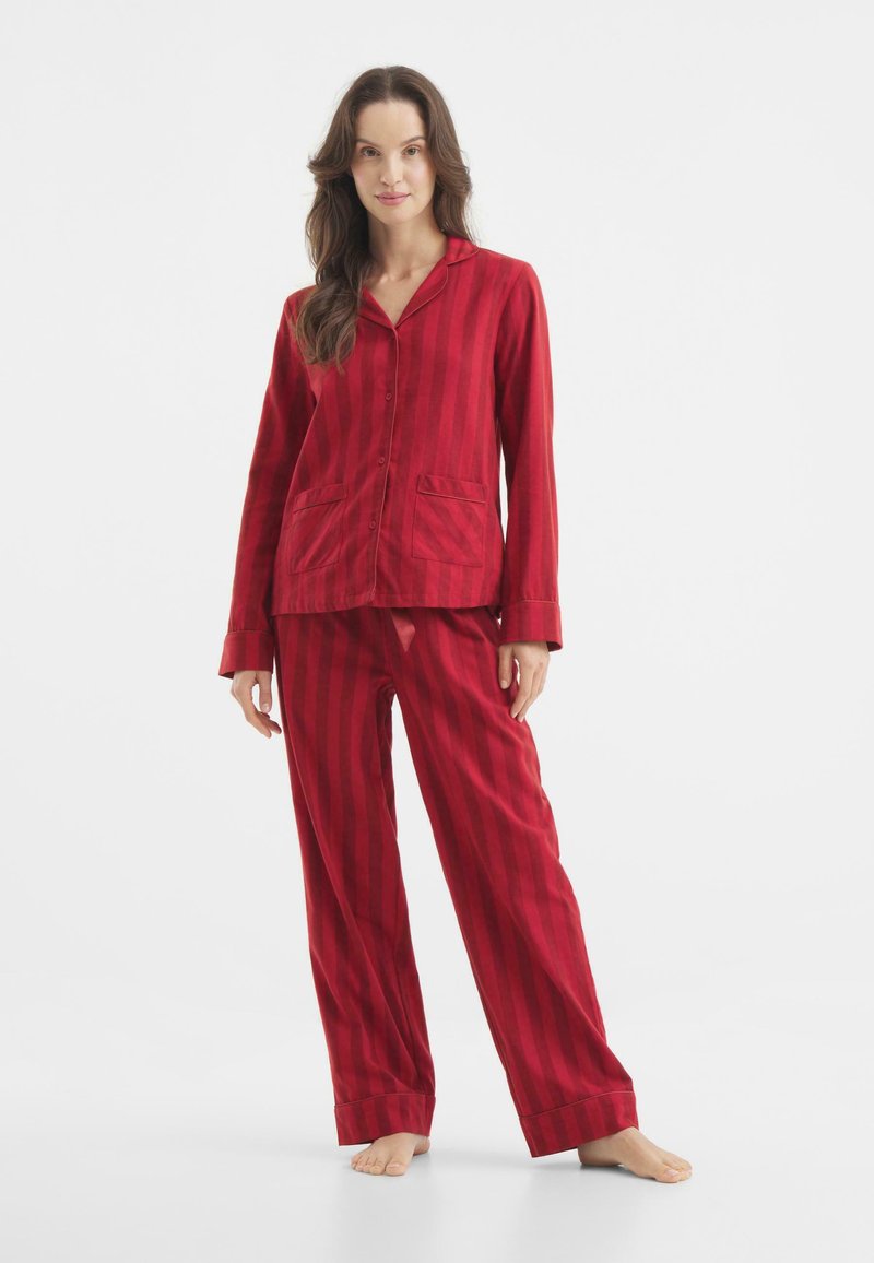 Punainen raidallinen pyjama-setti, jossa on napitettavat paita ja vastaavat housut. Kangas näyttää pehmeältä ja siinä on klassinen kaulus sekä kaksi etutaskua.