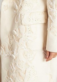 Abrigo blanco de lana con intrincados bordados florales en cuentas doradas y acentos en hilo blanco, grandes botones y un bolsillo de parche.