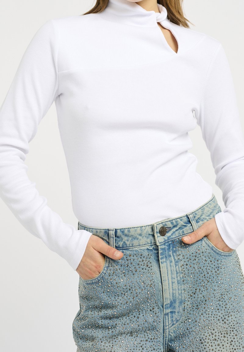 Femme portant un haut blanc côtelé à manches longues avec une petite découpe au niveau du cou et un jean bleu clair taille haute orné de strass.