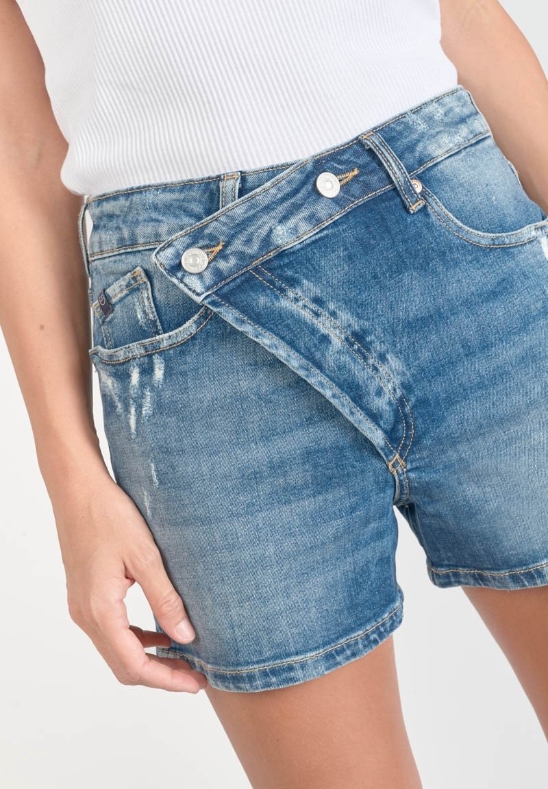 Femme portant un short en jean bleu avec un rabat asymétrique boutonné à l'avant et un haut blanc côtelé.