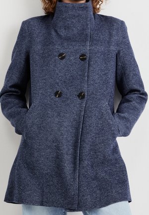 Femme portant un manteau en laine bleu marine à double boutonnage avec des boutons noirs, debout les mains dans les poches devant un fond uni.
