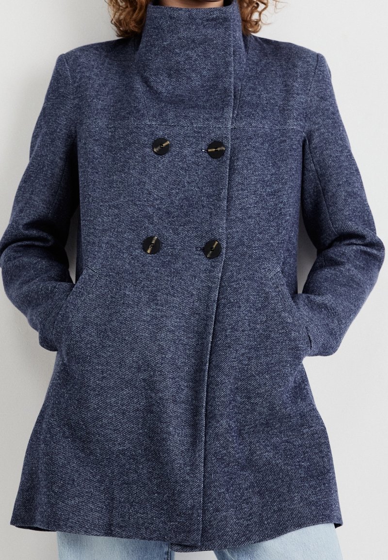 Femme portant un manteau en laine bleu marine à double boutonnage avec des boutons noirs, debout les mains dans les poches devant un fond uni.