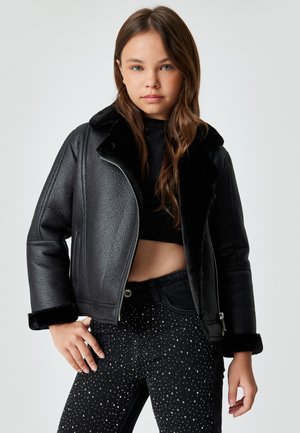 Jonge vrouw in een zwarte cropped shearling jas, zwarte crop top en zwart met studs versierde broek, staande tegen een effen achtergrond.