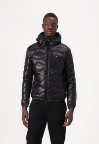 Giacca a vento nera con finitura lucida, dotata di cappuccio, chiusura frontale con zip e una piccola tasca sul petto. Accentuata con rifiniture verdi.