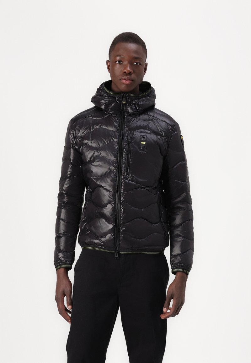 Giacca a vento nera con finitura lucida, dotata di cappuccio, chiusura frontale con zip e una piccola tasca sul petto. Accentuata con rifiniture verdi.