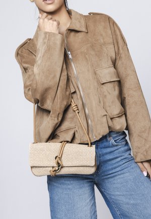 Ženska, oblečena v rjavo antilop jakno in modre kavbojke, nosi bež prepleteno torbico čez ramo z usnjenim trakom s vozlom.