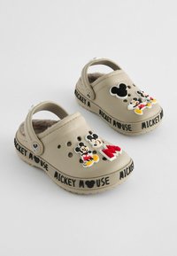 Beige tupsut, joissa on pehmeä keinoturkki vuori ja joissa on erilaisia värikkäitä Mikki Hiiri -kuvioita sekä musta "MICKEY MOUSE" -teksti sivuilla.