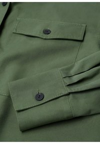 Manche de chemise en tissu vert et poche poitrine avec boutons noirs et détails de couture visibles.