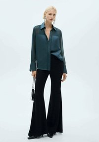 Teal sheer button-up shirt with long sleeves, paired with high-waisted black flared pants. Model holds a small black handbag.