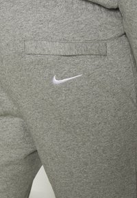 Närbild av grå sweatpants med ett bakfack och en vit Nike swoosh-logotyp broderad under fickan.