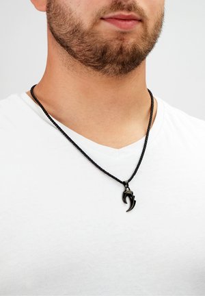 Black woven necklace featuring a claw-shaped pendant. The pendant is smooth with a glossy finish. Worn over a white shirt.