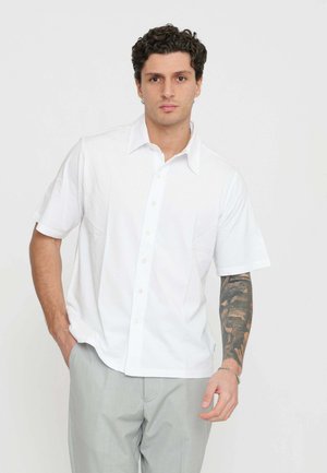 Camicia bianca a maniche corte con colletto e vestibilità rilassata. Tessuto morbido, abbinata a pantaloni grigio chiaro.