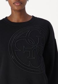 Sudadera negra con un diseño de logo en relieve tonal en el centro, cuello redondo y tejido texturizado que aporta profundidad.