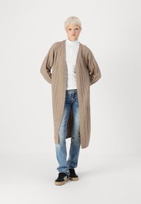 Pikk beeži soonikkardigan avatud ette, kombineeritud valge kõrge kraega pluusiga ja tuhmide siniste teksadega. Mustad tossud beežide talladega.