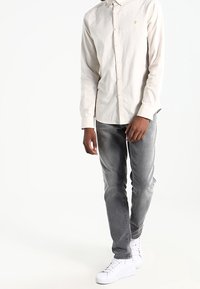 Hellbeiges Hemd zum Knöpfen mit langen Ärmeln und kleinem Logo. Kombiniert mit grauer Jeans und weißen Sneakern. Einfaches, lässiges Design.