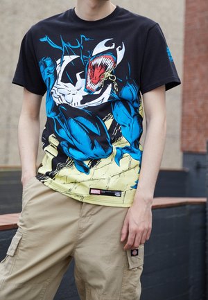 Person wearing a black Marvel T-shirt with a large blue and white venom graphic, paired with beige cargo pants.