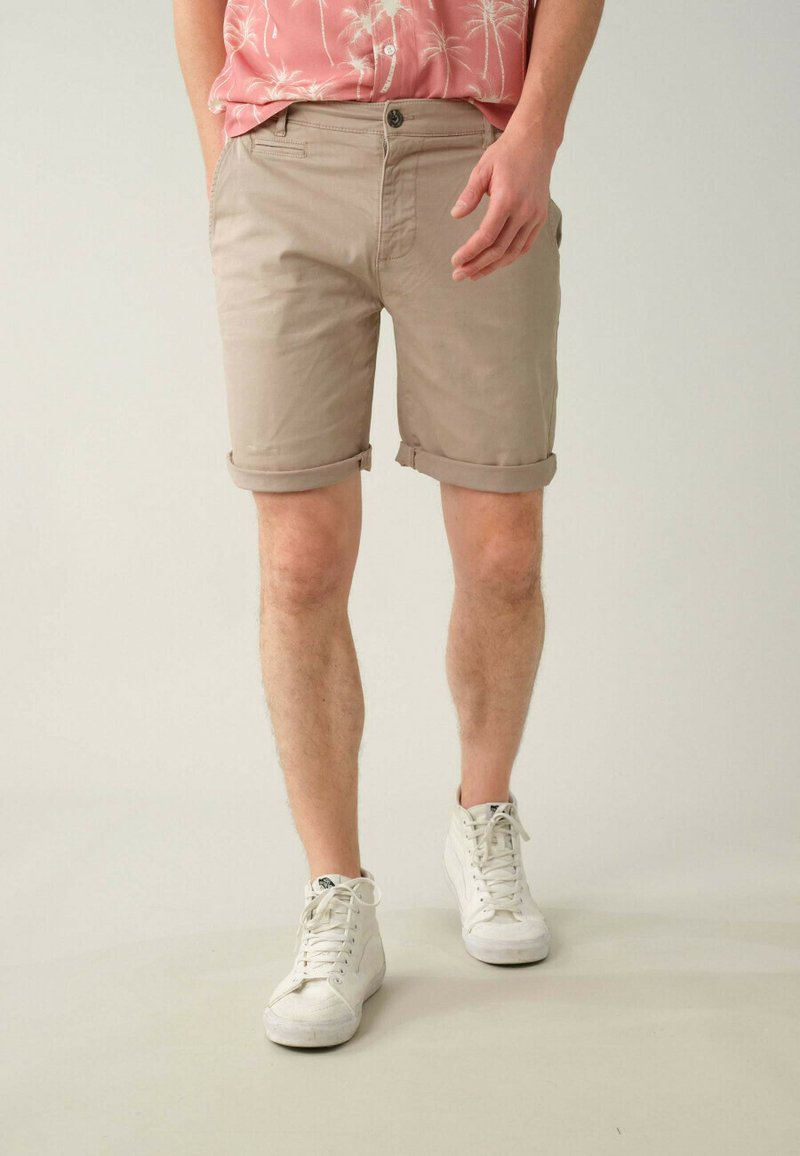 Shorts en coton beige avec revers, dotés d'une poche avant et portés avec des baskets blanches. Design décontracté pour les temps chauds.