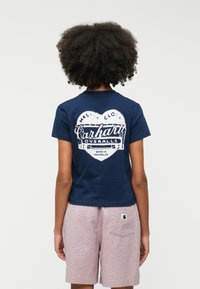 Mørkeblå t-shirt med hvid grafisk print på ryggen, der viser "Carhartt Overalls." Sættet er kombineret med lyserøde teksturerede shorts.