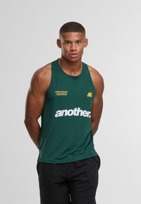 Svalnatý muž s krátkými vlasy v zeleném sportovním tílku s nápisem "další", stojící s rukama za zády na jednobarevném pozadí.