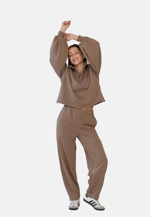 Kobieta w brązowej pasującej bluzie i spodniach, rozciągająca się z uniesionymi ramionami, uśmiechająca się, ubrana w białe i czarne trampki, stojąca na jednolitym tle.