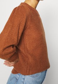 Persoon in een gestructureerde roestkleurige gebreide trui met ballonsleeves en lichtblauwe denim jeans, staand in zijprofiel met de handen achter de rug.
