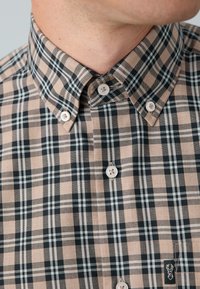 Chemise à manches courtes à carreaux beige et noir, dotée d'un col boutonné, de boutons blancs et d'un petit emblème à l'ourlet.